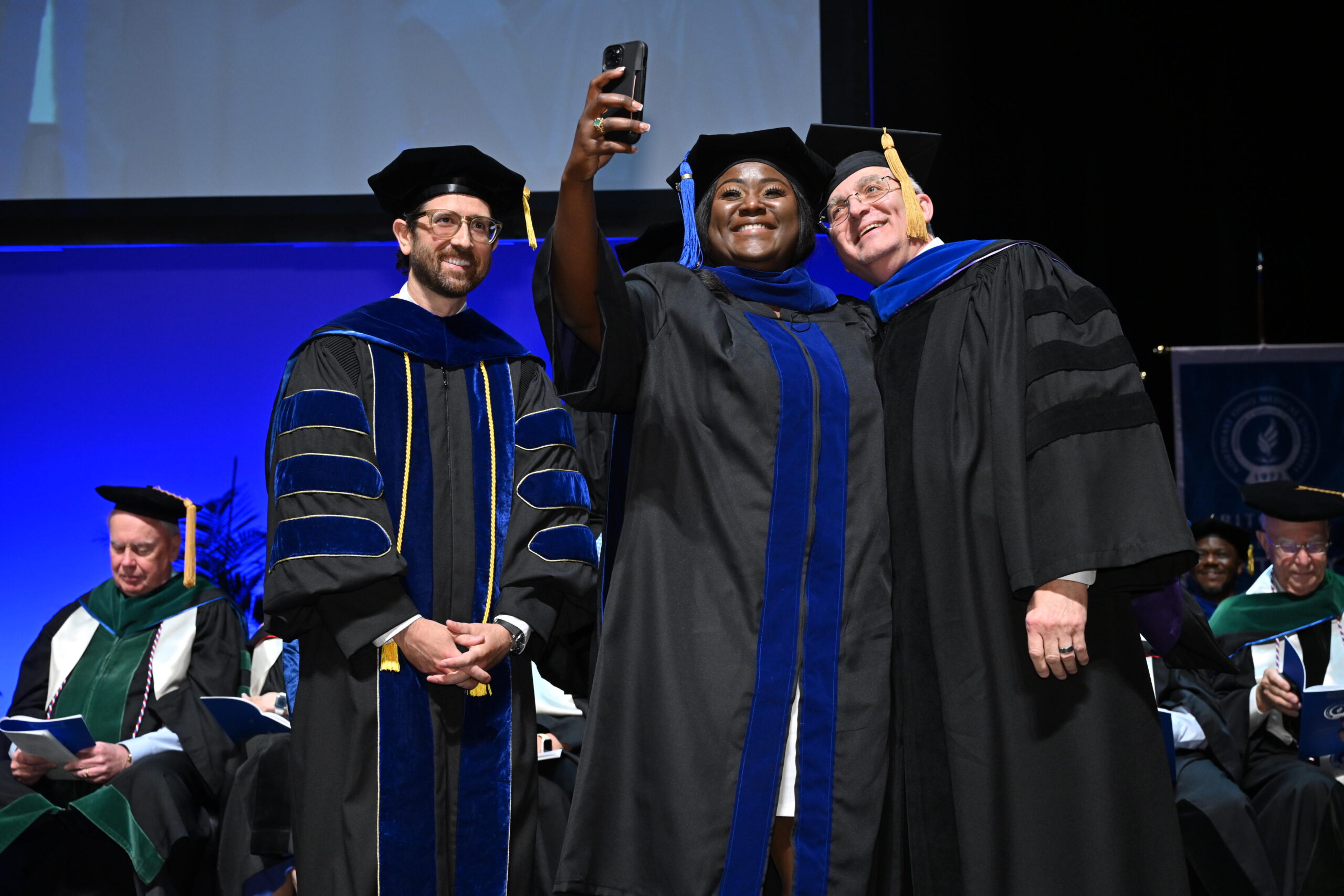 A woman and two men dressed in academic regalia pose for a selfie on stage at a graduation ceremony