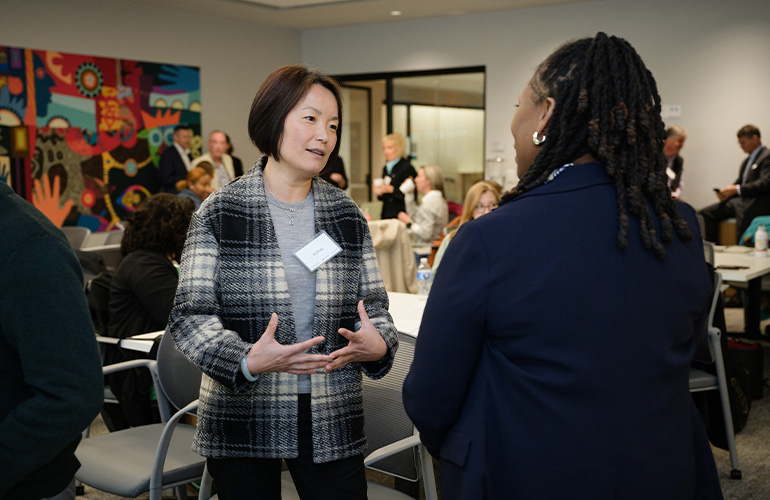 woman in black and white plaid sweater gestures with her hands while talking to another women whose back is to the camera