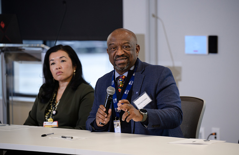 A man and a woman seated at a table while participating in a panel discussion. The man speaks into a handheld microphone