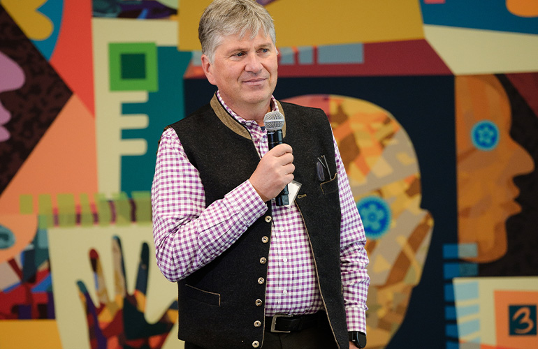 A man in button shirt and vest stands in front of a colorful backdrop while speaking into a microphone