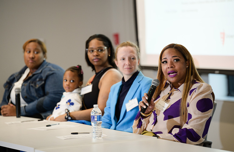 Four women seated at a table during a panel discussion. One woman speaks into a microphone. Another one holds a child on her lap.