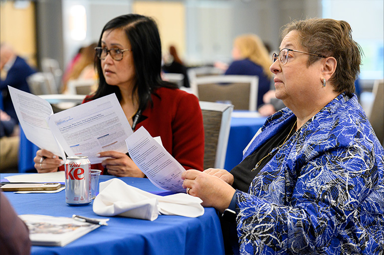 two women in business attire are seated a table. They hold papers in their hands and look across the table with serious expressions
