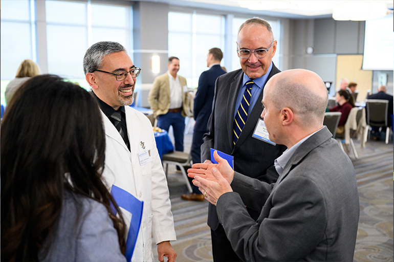 two men in suits chat with a man in a white coat. a woman with her back to the camera joins in their conversation