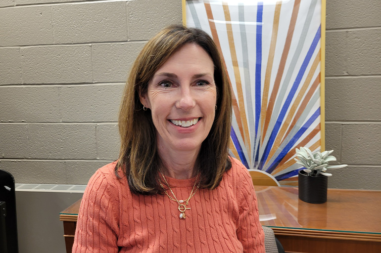 a woman smiles while seated in front of a colorful piece of artwork