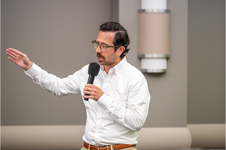 A man in a white shirt speaks into a microphone and gestures with his right hand