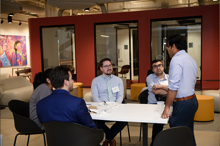 a man stands and addresses people seated around a table
