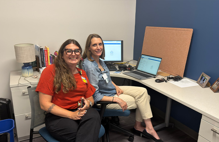 Two women smile at the camera while seated in an office