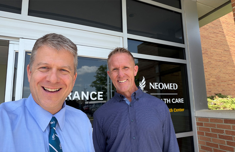 two men in blue shirts pose for a selfie outside a glass door with the NEOMED logo in the background
