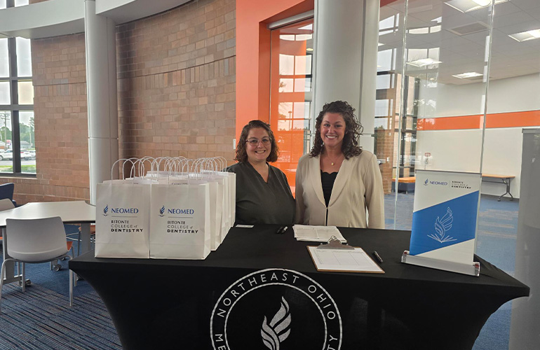 Two women stand behind a welcome table with a NEOMED tablecloth on it. Several bags branded with the logo of the Bitonte College of Dentistry sit on one side of the table. On the other side is a small pop-up banner with College branding on it
