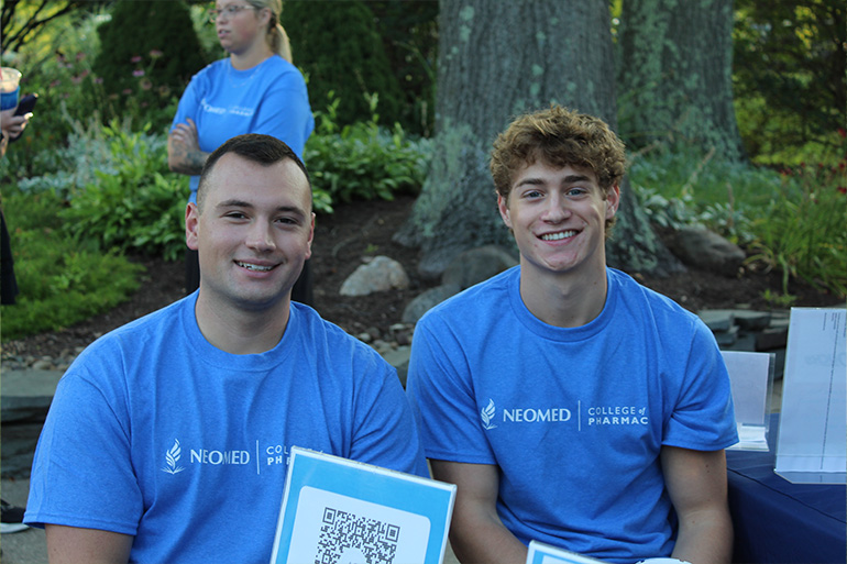 two young men in blue T-shirts that include the NEOMED College of Pharmacy logo in white sit a table