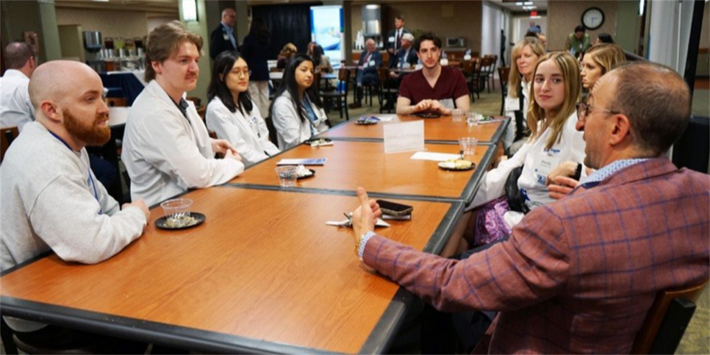 A group of health professions students in white coats are seated at a table talking to a man in a suit