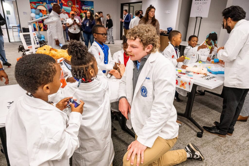 A man in white coat kneels while talking to a group in children in white coats