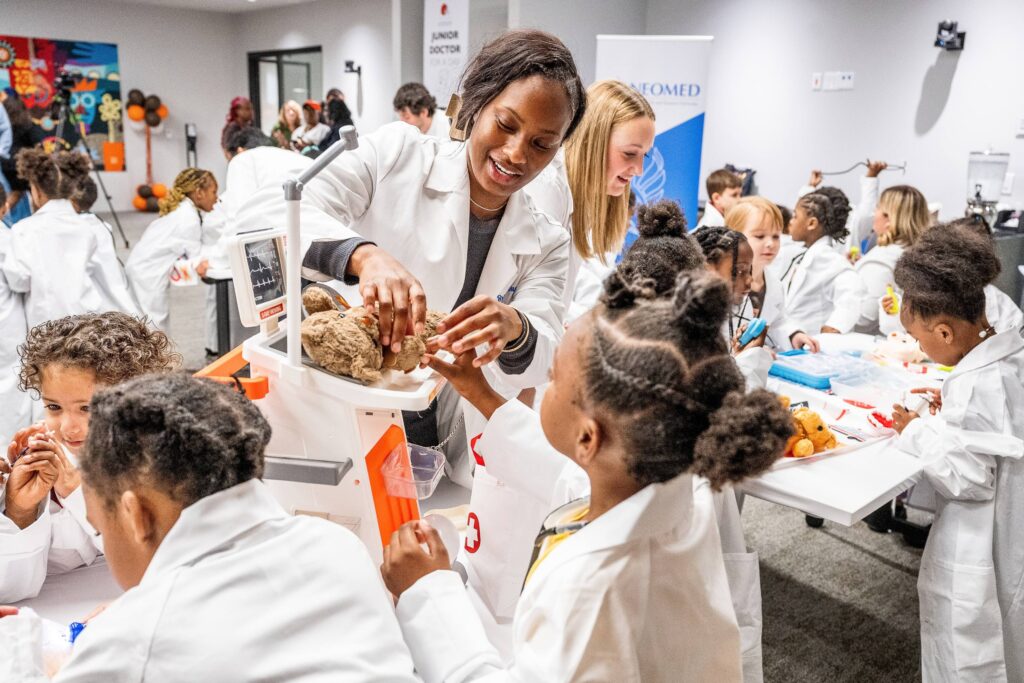 A woman in white coat helps a child in white coat weigh a teddy bear during a 'medical exam'