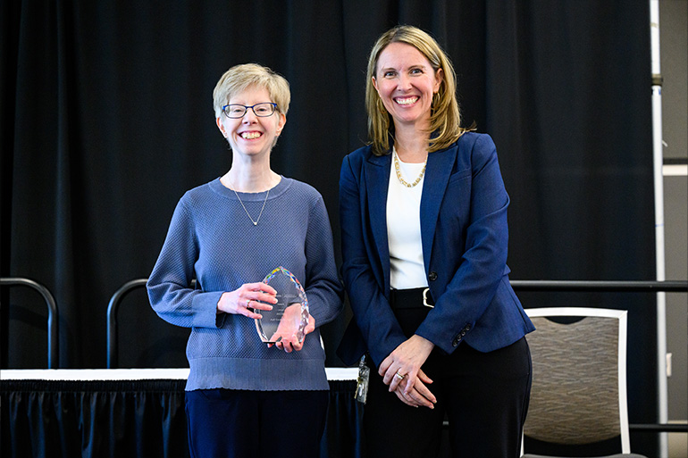Two woman pose on stage. The woman on the left holds a glass award
