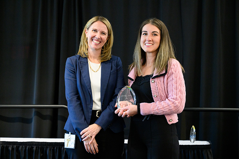 Two woman pose on a stage. The woman on the right holds a glass award