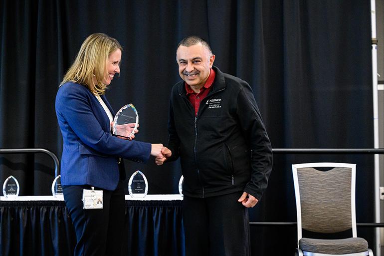 A woman in a blue jacket shakes the hand of a man in a branded trainer. She is about to hand him a glass award