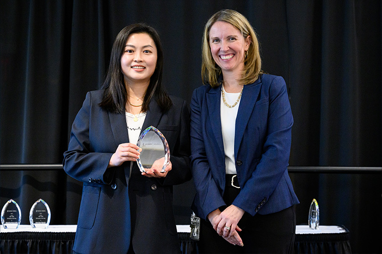 Two woman in business attire pose on a stage. One woman holds a glass award