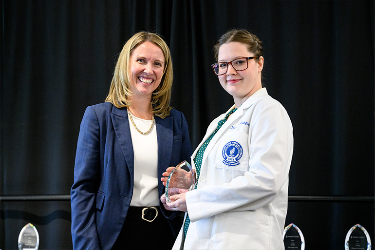 Two woman pose on a stage. The woman on the right is wearing a white coat and holding a glass award