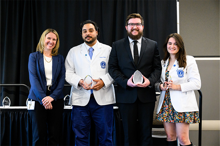 Four people, two in white coats, pose in a line. Three of them hold glass awards