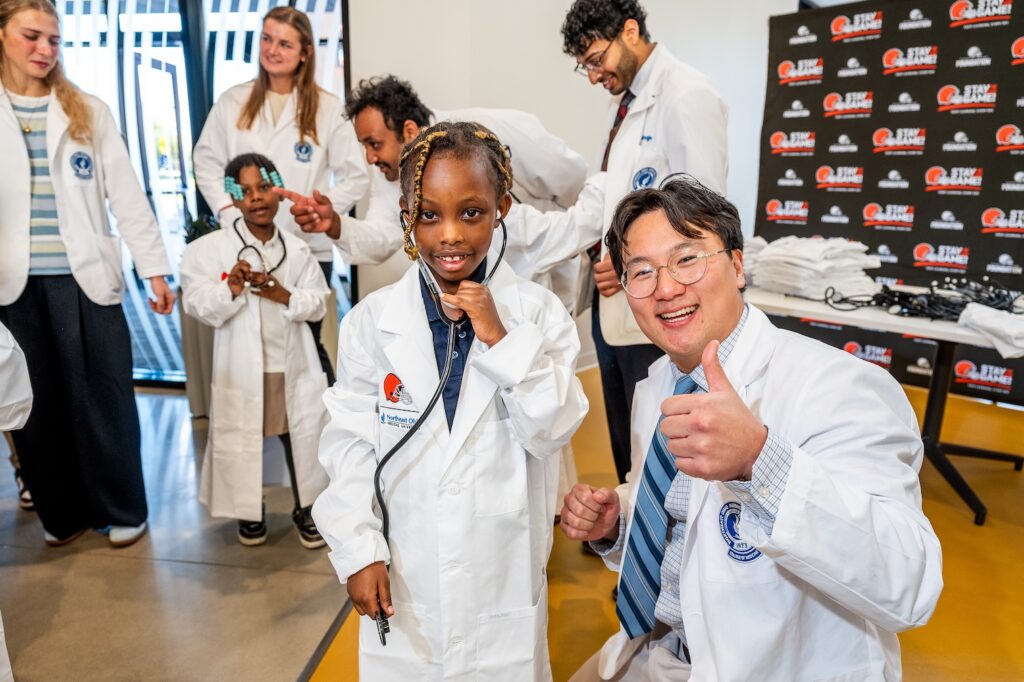 A man in white coat gives a thumbs up while posing with a child in a white coat with a stethoscope
