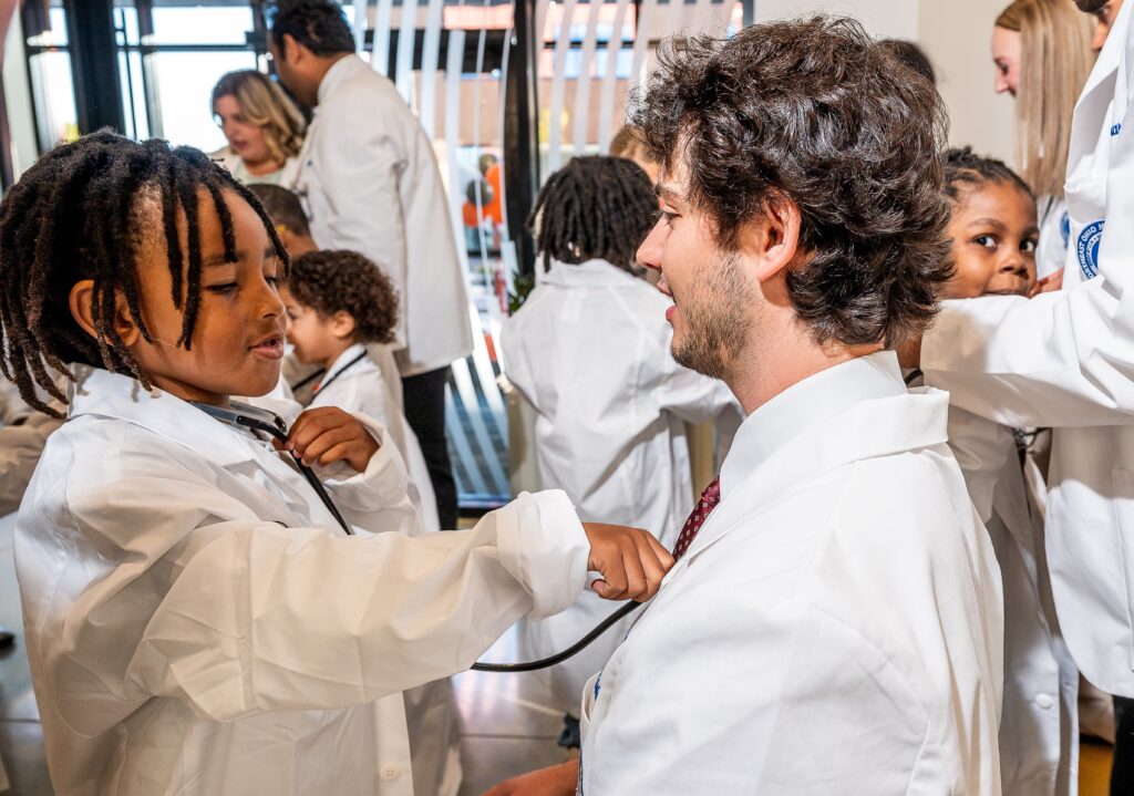 A child in a white coat uses a stethoscope to listen to the heartbeat of a man in a white coat
