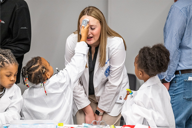 A child holds a thermometer to the forehead of woman in a white coat while other children look on