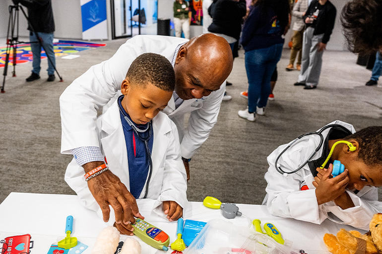 A man in white coat helps a little boy, also in white coat, with a toy medical device