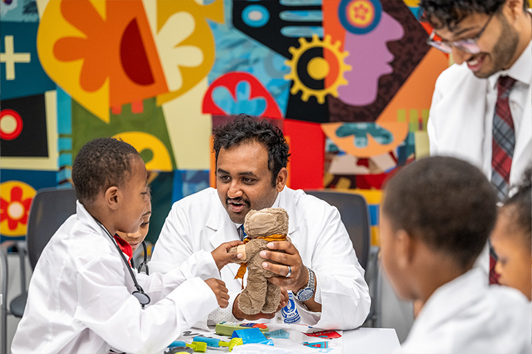 A man in white coat holds a teddy bear as a child in white coat pretends to listen for the bear's heart beat
