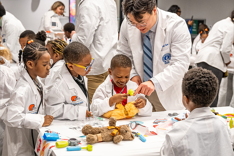 A man in white coat helps three children in white coats check the ears of a teddy bear