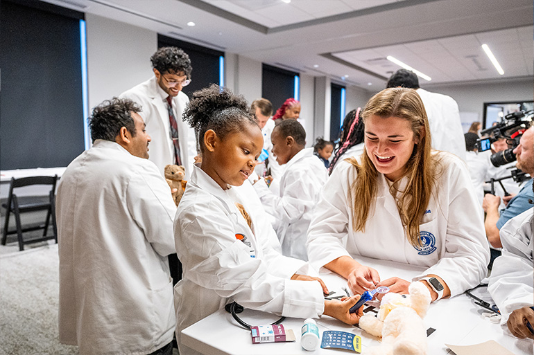 A little girl with white coat pretends to give medicine to a teddy bear while a smiling woman in white coat looks on