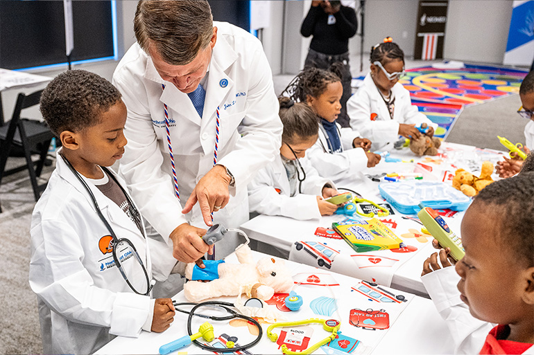 A man in a white coat shows a toy medical device to a child in a white coat