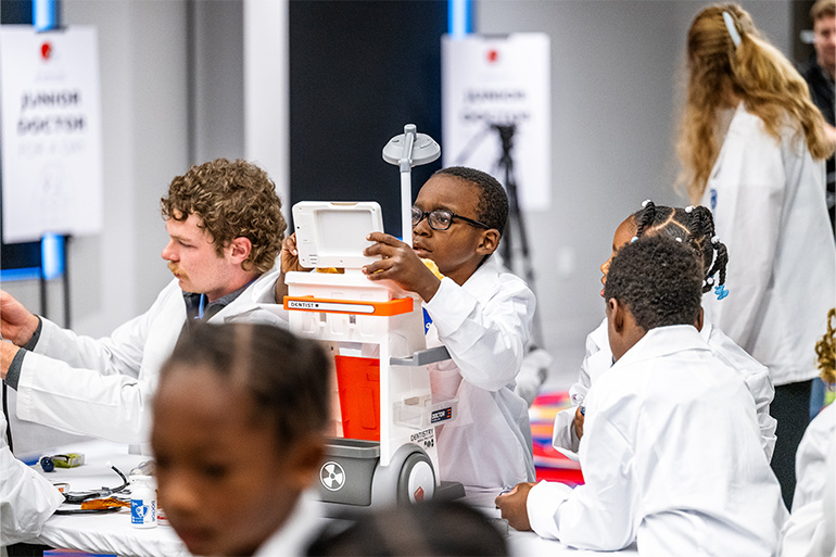 a child in a white coat looks very seriously at a scale in the center of a large group of children and adults in white coats