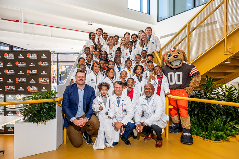 A group of children and adults in white coats pose on as stairway with the Browns mascot and team physician