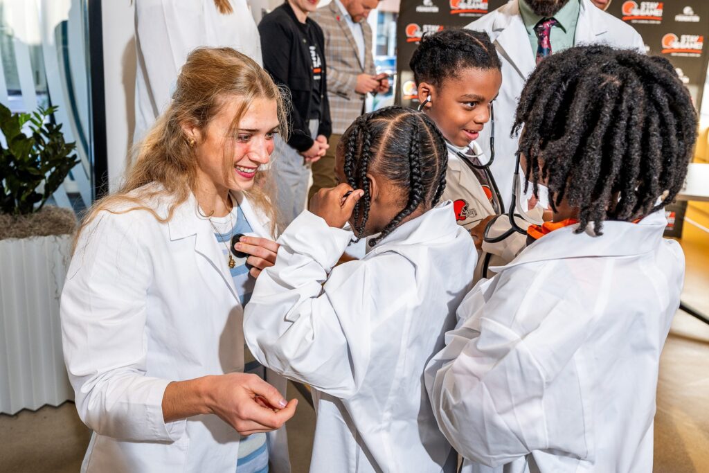 A woman in white coat helps a child in white coat adjust a stethoscope on their ears