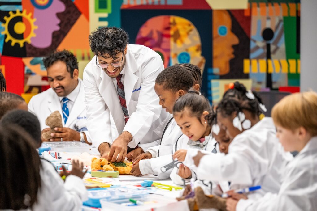 A man in white coat demonstrates how to use a stethoscope on a teddy bear to a child in white coat